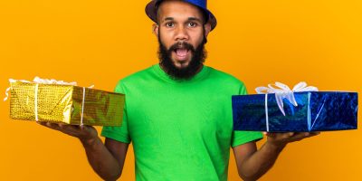 surprised young afro-american guy wearing party hat holding gift boxes isolated on orange background